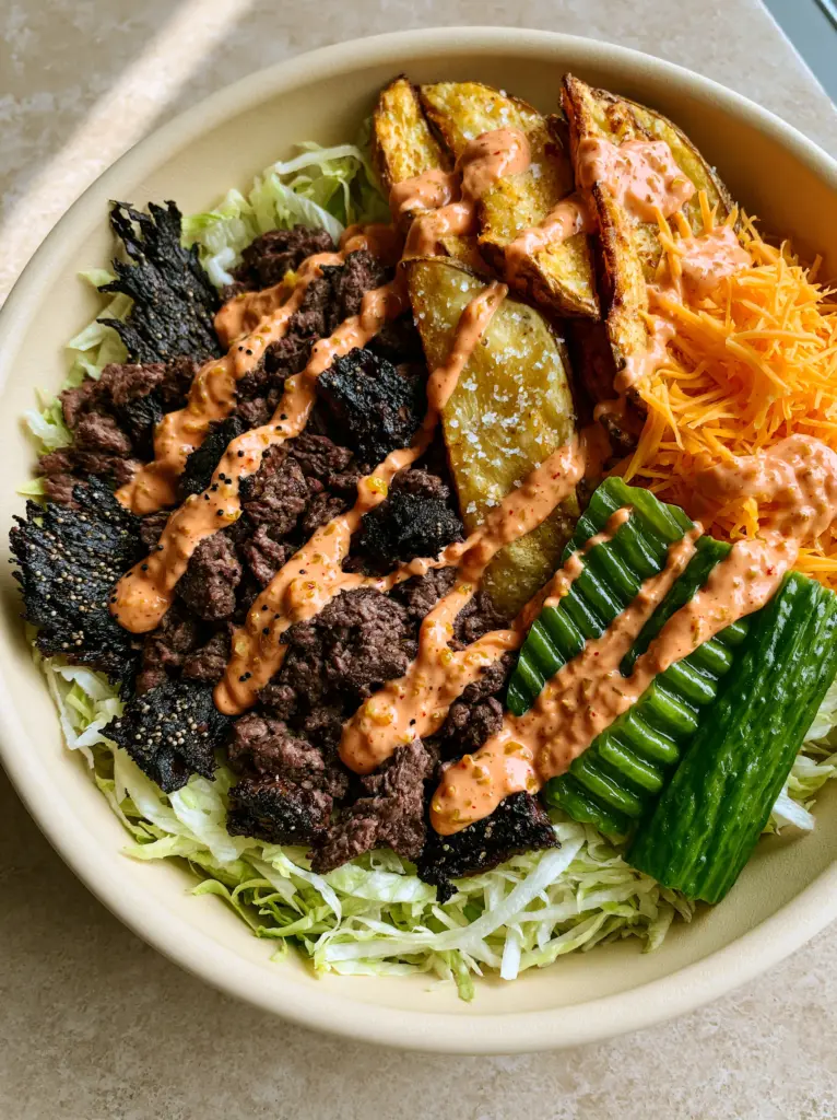 A handheld photo of a Burger Bowl with artistic drizzle of orange special sauce and green pickles.