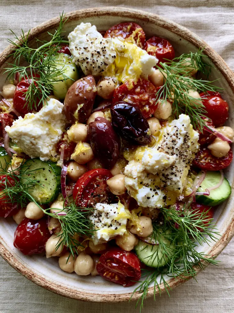 Greek Chickpea Salad with Herb-Infused Olive Oil and Rustic Feta 1 A rustic handcrafted ceramic bowl containing greek chickpea salad garnished with glossy Kalamata olives and hand-crumbled feta chunks.