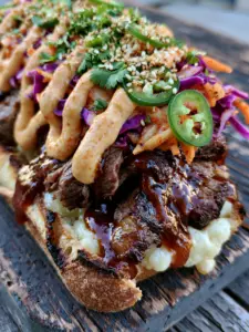 Extreme close-up of seared ribeye strips with a dark amber bulgogi glaze and melted mozzarella on a charred baguette. (Korean Steak Sandwich)