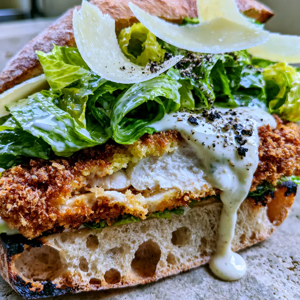 A close-up of the Chicken Caesar Sandwich featuring shaved parmesan curls and cracked black pepper.